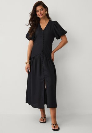 Kvinde stående iført en sort midi kjole med pufærmer, knapper foran, sideslidsel og sorte thongsandaler mod en ensartet lys baggrund.