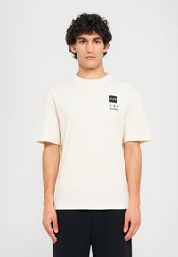Cream-colored cotton t-shirt with short sleeves, crew neckline, and a black logo patch on the left chest area. Soft texture.