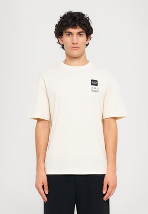 Cream-colored cotton t-shirt with short sleeves, crew neckline, and a black logo patch on the left chest area. Soft texture.
