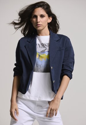 Navy blue corduroy blazer with rolled sleeves, silver buttons, layered over a white graphic t-shirt and white pants. Smooth texture.