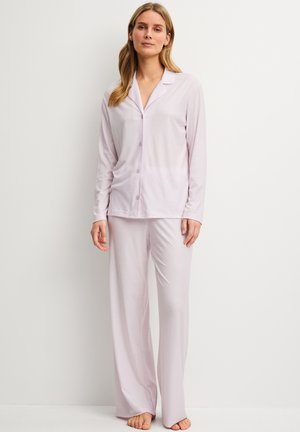 Femme aux longs cheveux portant un haut rayé rose clair à manches longues boutonné et un pantalon large assorti, debout pieds nus contre un mur blanc.