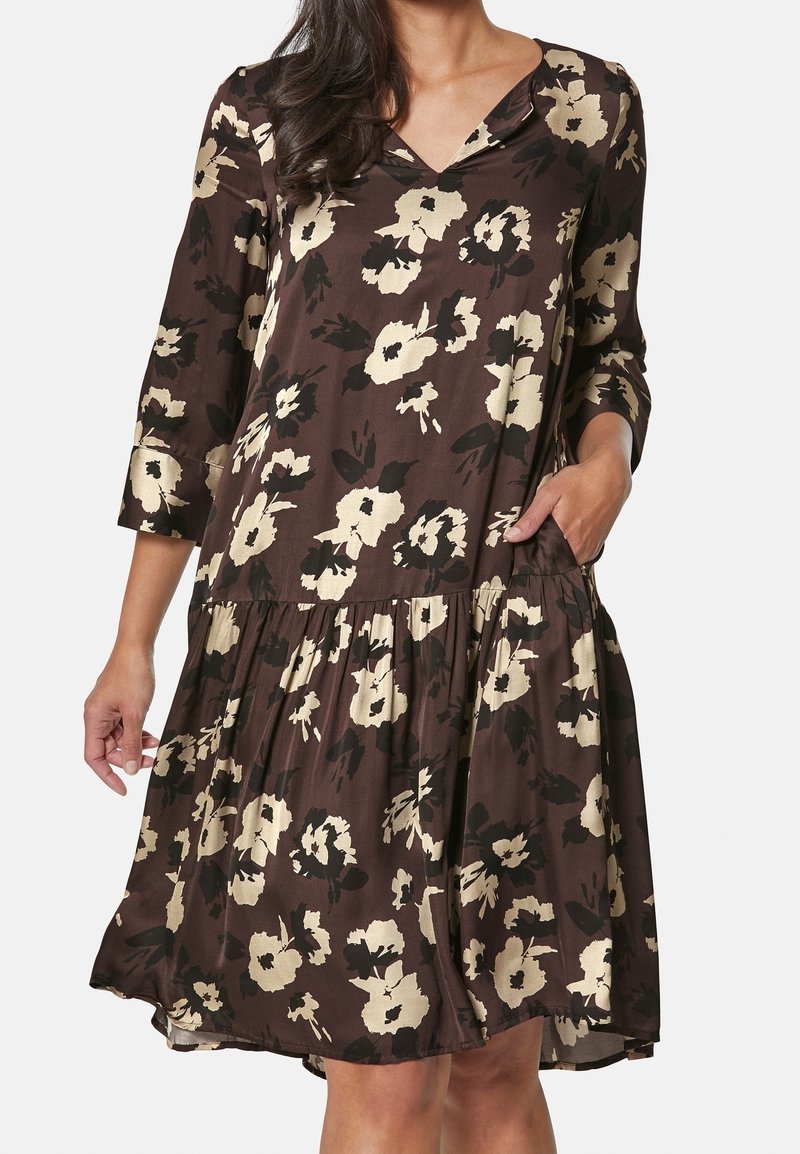 Femme portant une robe marron avec des motifs floraux crème et noirs, manches trois-quarts et une jupe ample plissée, main reposant dans une poche latérale.