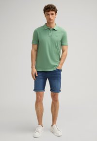 Polo verde con una texture morbida, abbinato a pantaloni shorts in denim arrotolati e sneakers bianche. Dettaglio del logo ben visibile sulla maglietta.