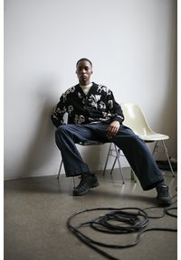 Mladić u crnom kardiganu s uzorkom i tamnoplavim hlačama sjedi raširenih nogu na stolici u minimalističkoj sobi s namotanim kabelom na podu.