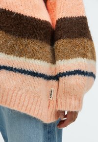 Chunky knit sweater in pastel hues of pink, brown, and cream. Features a ribbed cuff and a label with "SENSES" on the hem.