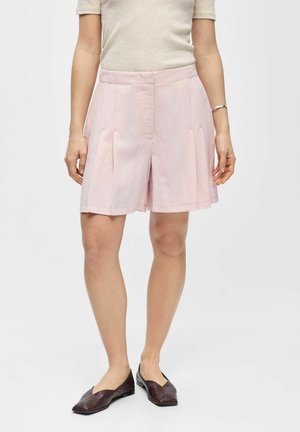 Kvinne iført lys rosa plissert shorts, en beige topp med korte ermer, mørkebrune slip-on sko og et sølvarmt armbånd på venstre håndledd.