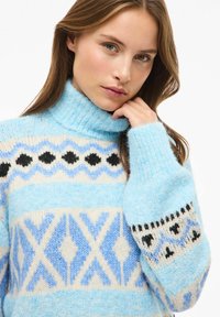 Hellblauer Strickpullover mit hohem Kragen, mit geometrischen Mustern in Weiß und dunkleren Blautönen, akzentuiert mit schwarzen Designs an den Ärmeln.