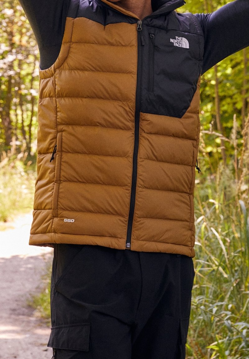Tans- und schwarzes Steppwesten mit gesteppten Bereichen, mit Reißverschluss vorne und Logo auf der Brust. Über einem dunklen Outfit getragen, in einer natürlichen Umgebung im Freien.