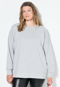 Mladá žena s vlnitými vlasy má na sobě světle šedou oversize mikinu, černé kožené kalhoty a zelený oční stín, stojí proti bílému pozadí.