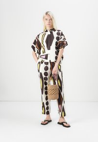 Ensemble deux pièces comprenant une chemise à manches courtes à motifs et un pantalon de couleur blanche, brune, jaune et violette. Comprend un sac en osier et des tongs.