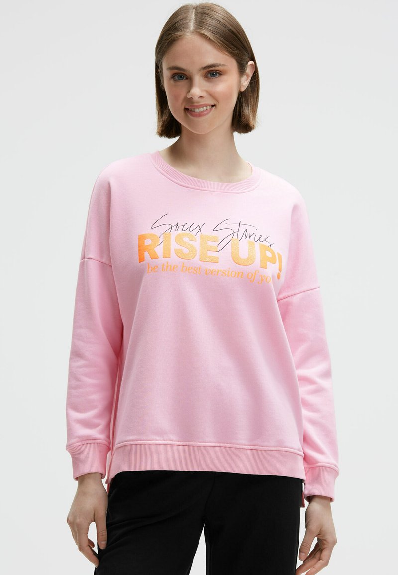 Junge Frau in einem rosa Sweatshirt mit dem Aufdruck "Sioux Stories RISE UP! Sei die beste Version von dir selbst", lächelt vor einem schlichten Hintergrund.
