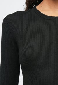 Žena u uskoj crnoj rebrastoj dugoj majici s dugim rukavima i okruglim izrezom naspram jednostavne svijetle pozadine.