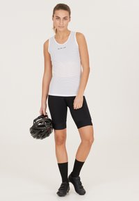 Hvid ærmeløs top med maskemønster, sorte cykelshorts, knæhøje sorte strømper, holder en sort cykelhjelm.