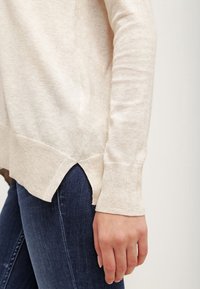 Pull beige en tricot avec manches longues, fentes latérales à l'ourlet, texture douce, porté avec un jean bleu foncé.