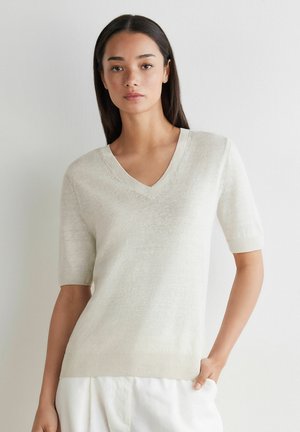 Ung kvinde iført en lysebeige V-hals kortærmet sweater og hvide bukser, stående mod en ensfarvet hvid baggrund.