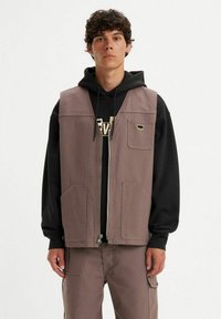 Giovane uomo che indossa un hoodie nero sotto un gilet marrone con zip e tre tasche, abbinato a pantaloni cargo.