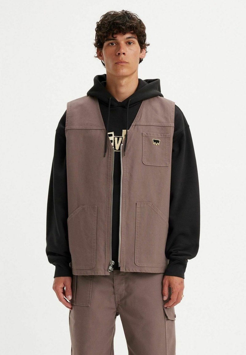 Giovane uomo che indossa un hoodie nero sotto un gilet marrone con zip e tre tasche, abbinato a pantaloni cargo.