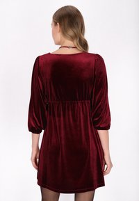 Rochie din catifea burgundy cu un croi lejer, manșete puff de 3/4 și talie cu volane, având o textură netedă și moale și o lungime până la genunchi.