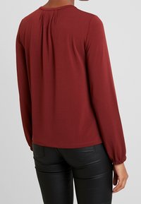 Bordowy top z długim rękawem i okrągłym dekoltem, z plisowaniem z tyłu i marszczonymi mankietami, w połączeniu z czarnymi dopasowanymi spodniami.
