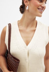 Cremfarbener ärmelloser Strickpullover mit V-Ausschnitt, gerippter Textur, versehen mit zwei Knöpfen, kombiniert mit einer gewebten braunen Tragetasche.