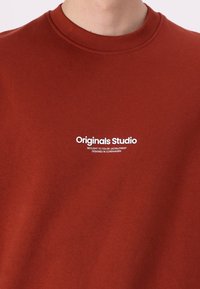 Roestkleurig sweatshirt met een ronde hals. Bevat witte tekst: "Originals Studio" met aanvullende details daaronder. Zachte katoenen textuur.