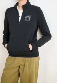 Person klädd i en svart Nike sweatshirt med kvartdragning, med händerna i framfickan och olivgröna byxor mot en neutral bakgrund.