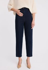 Kobieta ubrana w ciemnoniebieskie jeansy o wysokim stanie i prostych nogawkach, kremowy oversize'owy sweter, beżowe szpilki w szpic oraz dużą srebrną pierścionek na lewej ręce.