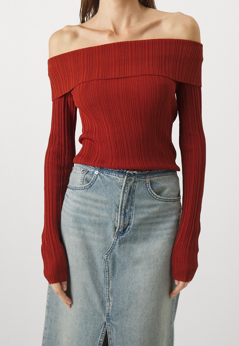 Femme portant un haut rouge côtelé à manches longues et épaules dénudées, associé à une jupe en denim bleu clair taille haute avec une fente sur le devant.