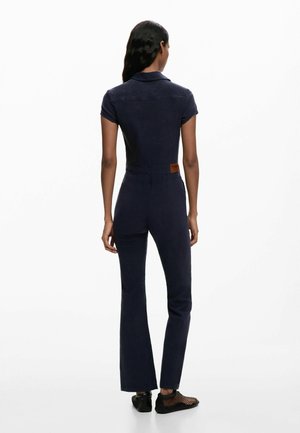 Hosszú fekete hajú nő, aki testhez álló tengerészkék overallt és fekete hálós tornacipőt visel, háttal állva, fehér háttéren.