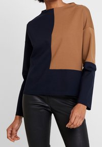 Camiseta de manga larga con un diseño en bloques de color en azul marino y marrón. El material es suave, con un ajuste relajado y un cuello redondeado alto.