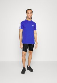 Blaues Sport-T-Shirt mit weißen Streifen an den Ärmeln, kombiniert mit schwarzen Shorts mit weißen Akzenten. Getragen mit schwarzen Sportschuhen.