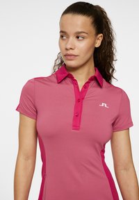 Tajtat passform rosa pikétröja med ribbad textur, korta ärmar och en treknappslåst. Har en kontrasterande färg på kragen och logotyp.