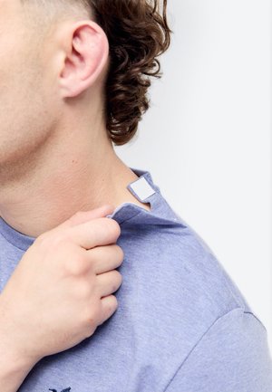 Personne aux cheveux bouclés tire sur le col d'une chemise bleu clair révélant une fermeture auto-agrippante blanche au niveau du cou pour un retrait facile.