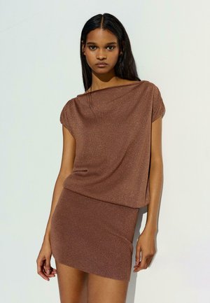 Mladá žena s dlhými tmavými vlasmi, oblečená v hnedom trblietavom tričku s krátkym rukávom a zodpovedajúcej priliehavej mini sukni, stojaca pred jednoduchým svetlým pozadím.