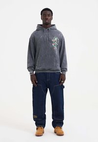 Grå hætte sweatshirt med blomsterbroderi, parret med blå denim cargo bukser. Brune arbejdssko fuldender outfittet.