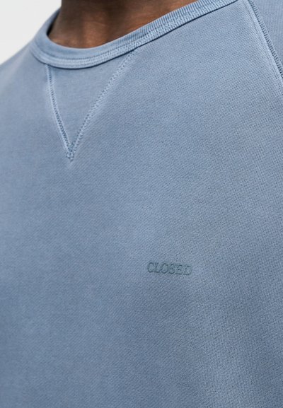 Personne portant un sweat-shirt bleu clair à col rond avec des détails cousus et le mot « CLOSED » brodé sur la poitrine.
