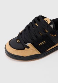 Baskets basses noir et beige avec de grosses lacets noirs, des panneaux en daim et en cuir, et le logo "GLOBE" sur le côté.