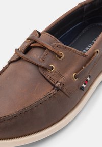 Zapato de barco de cuero marrón con ojales dorados, cordones planos y un acabado texturizado. Incluye una suela blanca contrastante y un pequeño detalle de logo.