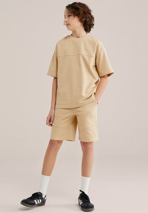 Lokkidega laps seisab käed taskutes, kandes beeži Blue Ridge t-särki ja lühikesi pükse, valgeid sokke ja musti tossusid.