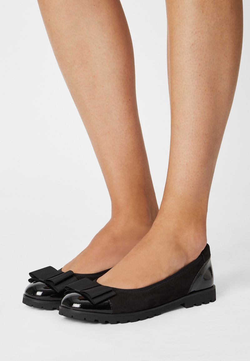 Pieds portant des chaussures plates noires avec des bouts brillants et de grands nœuds en tissu sur un fond blanc.