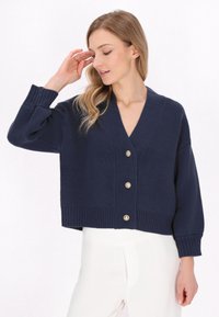 Cardigan in maglina di colore blu marino, con scollatura a V, maniche lunghe e orlo a costine, caratterizzato da cinque bottoni dorati sul davanti. Texture morbida.