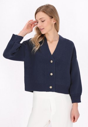 Cardigan tricotat de culoare bleu marin, cu decolteu în V, mâneci lungi și tiv nervurat, având cinci nasturi aurii pe față. Textură moale.