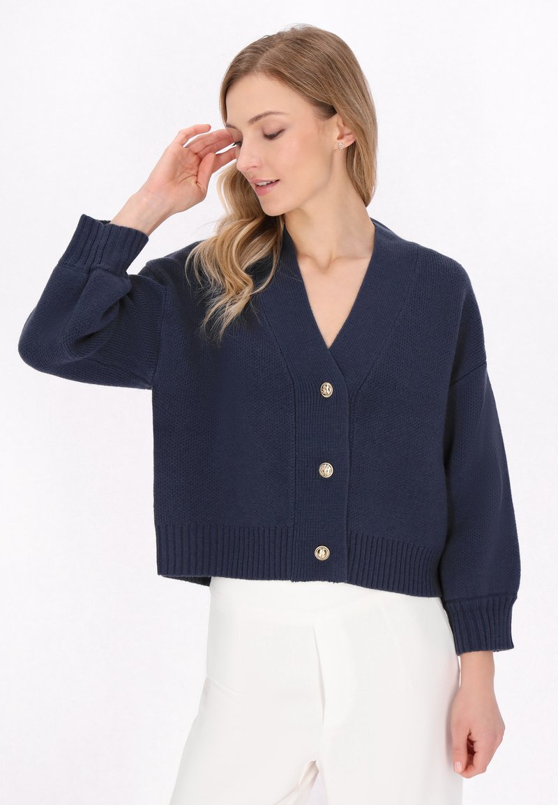 Cardigan in maglina di colore blu marino, con scollatura a V, maniche lunghe e orlo a costine, caratterizzato da cinque bottoni dorati sul davanti. Texture morbida.
