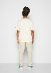 Krémové bavlněné tričko s volným střihem, krátkými rukávy a malým logem na levém rukávu, doplněné o světle béžové kalhoty a boty s zelenými akcenty.