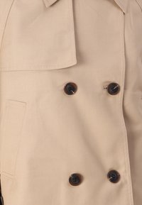 Cappotto trench beige doppiopetto realizzato in tessuto liscio, con grandi bottoni neri, colletto classico e tasche laterali.