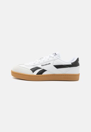 Zapatilla blanca con acentos negros, con una suela de goma texturizada, diseño de cordones y el logo de Reebok en el lateral y en la lengua.