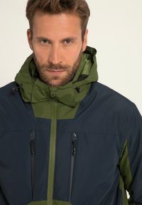 Herrejakke med tofarvet design i mørk marineblå og olivengrøn, tekstureret stof, justerbar hætte og lynlåslommer.