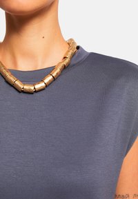 Szary top z krótkim rękawem ma gładką teksturę. Gruba złota naszyjnik z cylindrycznymi koralikami uzupełnia dekolt.