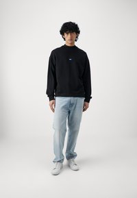 Zwarte sweatshirt met ronde hals en blauwe logodetails, gecombineerd met lichtblauwe jeans en witte sneakers op een model tegen een effen achtergrond.