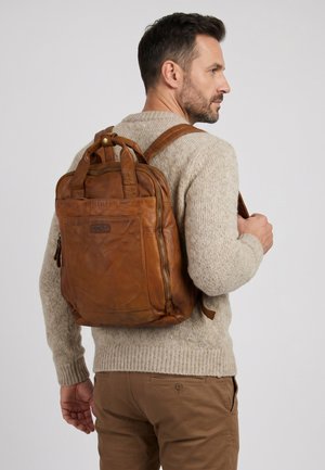 Mann mit beigem Pullover und brauner Hose trägt einen braunen Lederrucksack über einer Schulter und schaut seitlich vor einfarbigem Hintergrund.